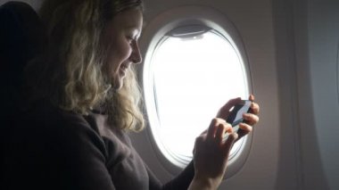 Kız uçak penceresinin yanına oturur ve telefondaki fotoğraflara bakar.