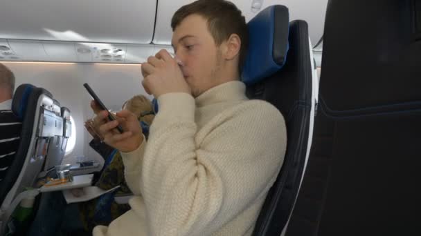 Mladý muž se dívá na telefon, zatímco sedí v letadle pije vodu — Stock video