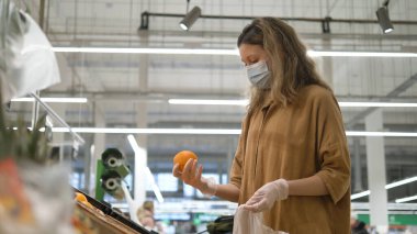 Tıbbi maskeli ve eldivenli bir kadın markette portakal seçiyor. Koronavirüs salgınından korunma, taze meyveye karşı bağışıklığın artması..