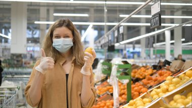 Tıbbi maskeli ve lastik eldivenli genç bir kadın marketten taze limon seçiyor, kameraya bakıyor, onaylıyor ve gülümsüyor. Koronavirüse karşı korunmak için artan bağışıklık.
