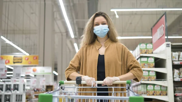 Vrouw met een medisch masker trekt beschermende rubberen handschoenen aan om zich te beschermen tegen het coronavirus in een supermarkt, neemt een boodschappenkarretje en gaat voor voedsel. — Stockfoto