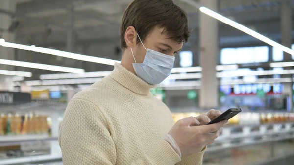 Man met medisch masker en rubberen handschoenen staat in een supermarkt met smartphone. Beschermende maatregelen tegen covid-19. — Stockfoto