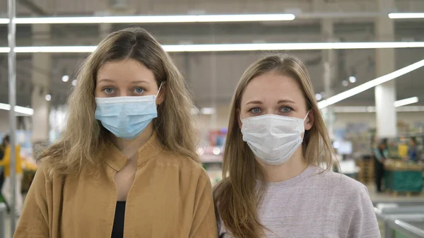 Две молодые женщины в медицинских масках стоят грустные в супермаркете и смотрят в камеру. Защита от пандемии коронавируса, подготовка к карантину . — стоковое фото