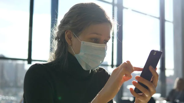 Tıbbi maskeli, ciddi yüzlü bir iş kadını akıllı telefonunun ekranına bakıyor, krizden ve koronavirüsten kaynaklanan mali kayıpları hesaplıyor.