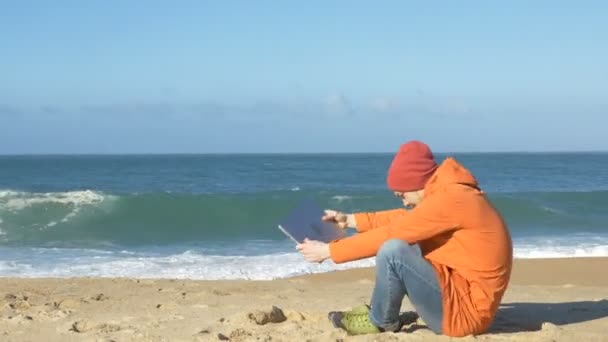 Uomo cammina lungo la spiaggia con il computer portatile si siede inizia a lavorare — Video Stock