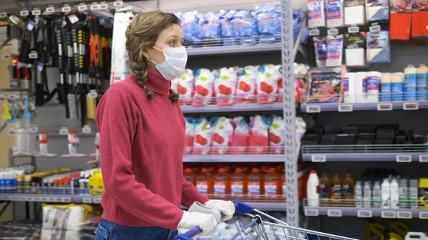 Girl in gloves and mask rolls food cart against background of goods in supermarket, protection coronavirus pandemic — Stock Photo, Image