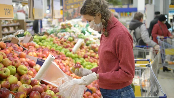 Tıbbi maskeli ve koruyucu eldivenli genç bir kadın süpermarkette elma topluyor. Koronavirüs pandemik koruması.