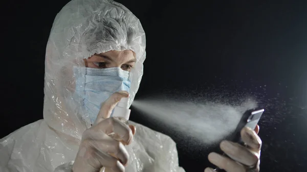 Людина в медичній масці і захисному костюмі розбризкує антисептик з мобільним телефоном на темному тлі. Захист коронавірусу. — стокове фото
