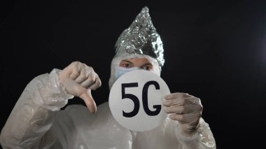 Koruyucu giysili, tıbbi maskeli ve folyo kapaklı bir adam 5 G işaretini kaldırıyor ve arka planda bir baş parmağı görünüyor. Kronavirüsün yayılması.