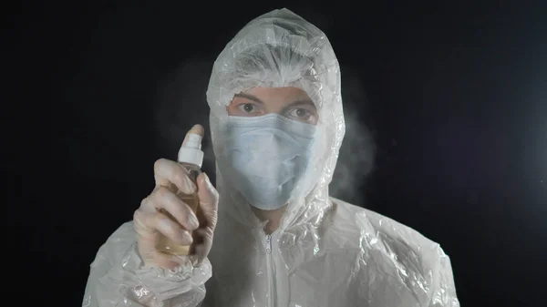 Портрет лікаря в медичній масці і захисний костюм розбризкує антисептичний спрей і дивиться на камеру на темному тлі — стокове фото