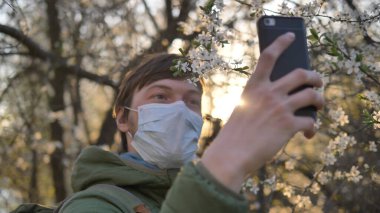 Coronavirus karantinası sırasında selfie, tıbbi maske takmış bir adam günbatımında akıllı telefonda çiçek açan elma ağacıyla fotoğraf çekiyor.