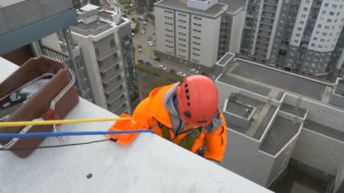 Turuncu takım elbiseli ve kasklı endüstriyel tırmanışçı cam yıkama için çok katlı binanın çatısından inmeye hazırlanıyor.