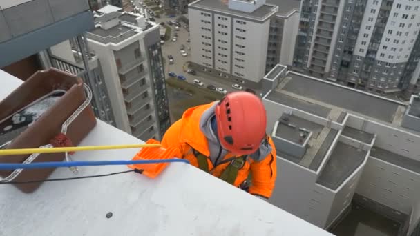 Industriklatrer i oransje dress og hjelm forbereder seg på å gå ned fra taket av fleretasjes bygning for glassvask – stockvideo