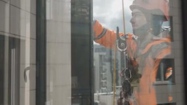 Gülümseyen endüstriyel tırmanışçı arka plandaki güneş ışığına karşı pencereleri yüksek irtifada yıkıyor
