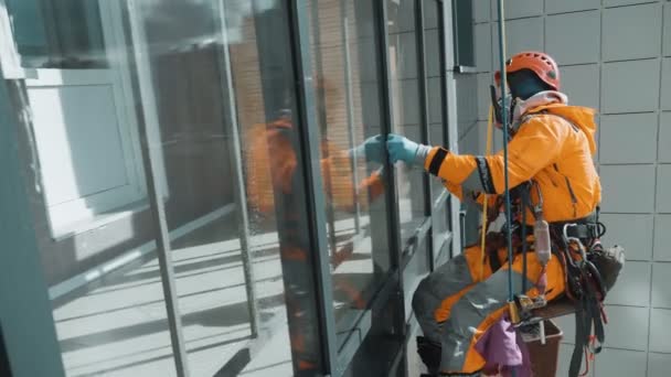 Стеклоочиститель, оборудованный промышленный альпинист в шлеме аккуратно моет окна снаружи здания — стоковое видео