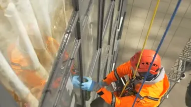 Miğferli ve lastik eldivenli bir cam temizleyicisi uzun bir binanın cephesini yıkıyor.