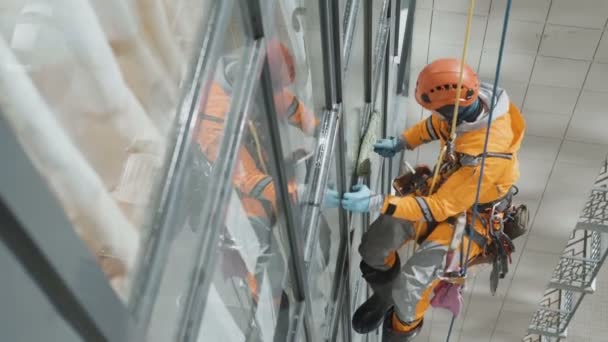 Miğferli endüstriyel tırmanışçı ve eldivenler yüksek binanın üst manzarasında camları siliyor — Stok video