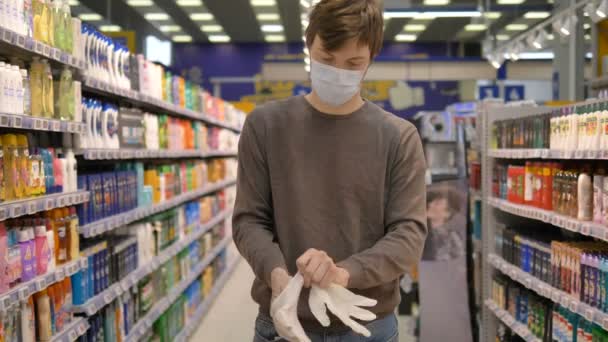 Coronavirus pandemische bescherming. Man met medisch masker doet rubberen handschoenen aan in een supermarkt tussen planken met goederen. — Stockvideo