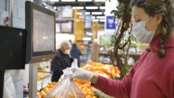 Egy nő orvosi maszkban és gumikesztyűben elektronikus mérlegen mérlegeli az árukat egy szupermarketben. A matrica a kesztyűhöz tapad.. — Stock videók