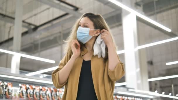 En ung flicka i en stormarknad sätter på sig en skyddande medicinsk mask och gummihandskar för att skydda mot infektion med coronavirus. — Stockvideo