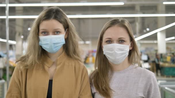 Tıp maskeli iki genç kadın bir süpermarkette üzgün üzgün dikiliyor ve kameraya bakıyor. Koronavirüs salgınından korunma, karantinaya hazırlık.. — Stok video