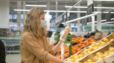 Tıbbi maskeli ve lastik eldivenli genç bir kadın marketten taze limon seçiyor, kameraya bakıyor, onaylıyor ve gülümsüyor. Koronavirüse karşı korunmak için artan bağışıklık.