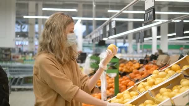 Uma jovem mulher em uma máscara médica e luvas de borracha seleciona limões frescos em um supermercado de supermercado, olha para a câmera, dá um polegar para cima e sorri. Aumento da imunidade para proteger contra o coronavírus . — Vídeo de Stock