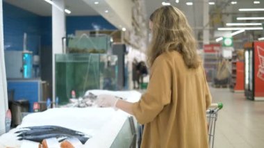 Süpermarkette tıbbi maske ve eldiven takmış genç bir kız balık satın alır. Koronavirüs enfeksiyonuna karşı önlemler.