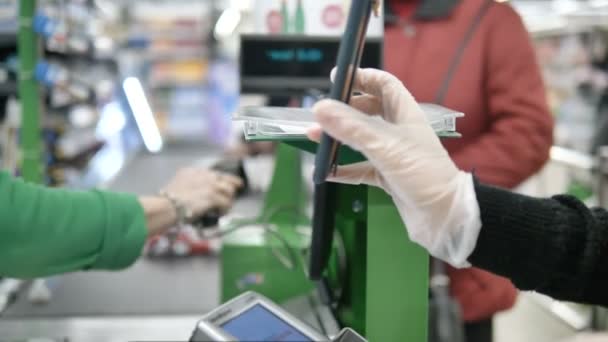 Una mujer en la caja en un supermercado le da a un cliente una tarjeta con un láser de su teléfono inteligente. Pagar en el supermercado con guantes protectores para no contraer el coronavirus — Vídeos de Stock