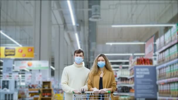 Pareja casada seria en una máscara médica y guantes de látex con carrito de comestibles está de pie en el supermercado, hay multitudes de clientes caminando, timelapse. Protección contra el coronavirus . — Vídeo de stock