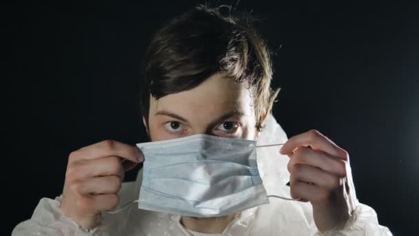 Koronavírus fertőzés. A fiatalember orvosi maszkot és fehér kezeslábast vesz fel csuklyával és hüvelykujjal felfelé. — Stock videók