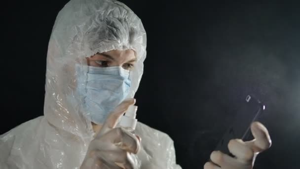Pria bertopeng medis dan jas pelindung menyemprotkan antiseptik dengan ponsel dengan latar belakang gelap. Perlindungan Coronavirus. — Stok Video