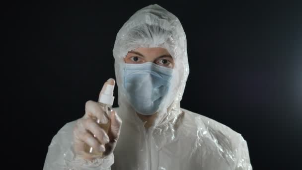 Πορτρέτο ενός γιατρού με ιατρική μάσκα και προστατευτική στολή ψεκάζει ένα αντισηπτικό σπρέι και κοιτάζει την κάμερα σε σκούρο φόντο — Αρχείο Βίντεο