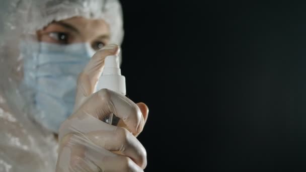 Coronavirus pandemie. Een arts in een medisch masker en beschermend pak spuit een antiseptische spray op een donkere achtergrond. Virusdesinfectie. — Stockvideo