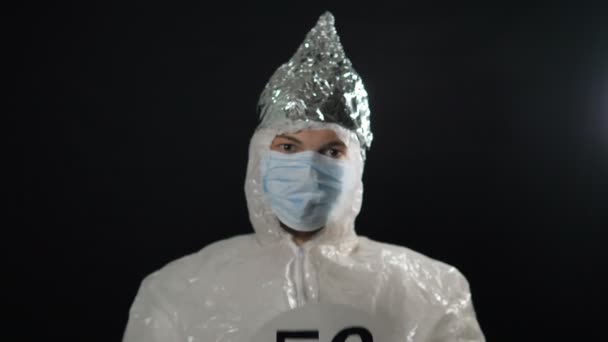 Mannen i skyddsdräkt, medicinsk mask och foliehätta höjer 5 g nätverksskylten och visar en tumme ner på en mörk bakgrund närbild. Spridning av kronavirus. — Stockvideo