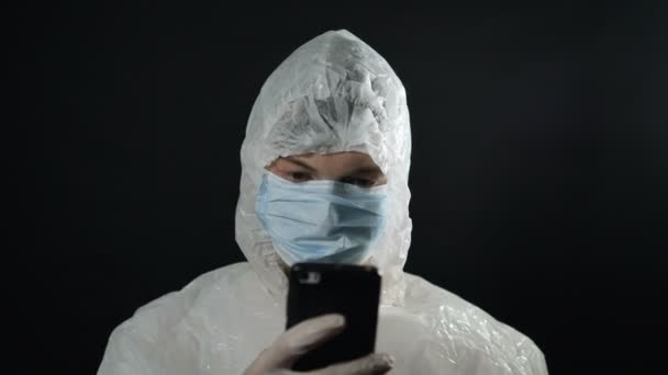 Homme en costume blanc et masque médical, met un capuchon en aluminium pour se protéger contre les radiations des tours 5G regarde smartphone sur fond noir close-up — Video