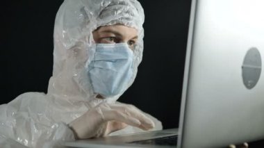 Tıbbi maskeli, lastik eldivenli ve koruyucu giysili bir virüs uzmanı karanlık bir arka planda dizüstü bilgisayarda çalışıyor. Koronavirüs salgınına karşı bir aşı geliştirilmesi.