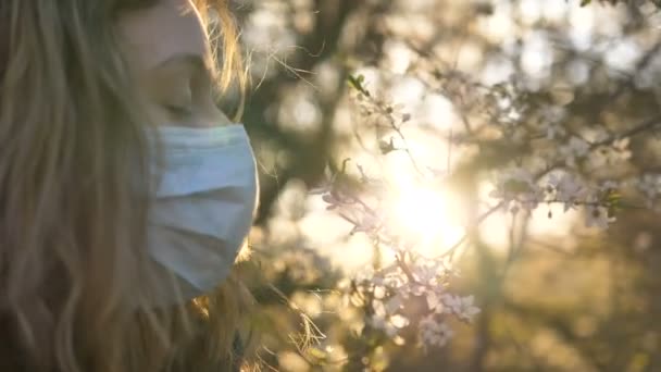 Fille dans un masque médical renifle un arbre à fleurs au coucher du soleil gros plan — Video