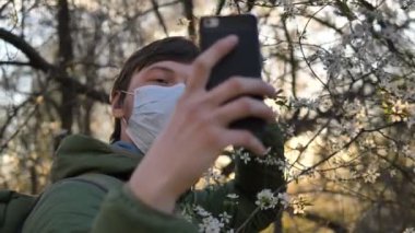 Tıp maskeli adam akıllı telefondan selfie çeker gün batımında çiçek açan elma ağacına, koronavirüs salgınına