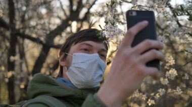 Coronavirus karantinası sırasında selfie, tıbbi maske takmış bir adam günbatımında akıllı telefonda çiçek açan elma ağacıyla fotoğraf çekiyor.