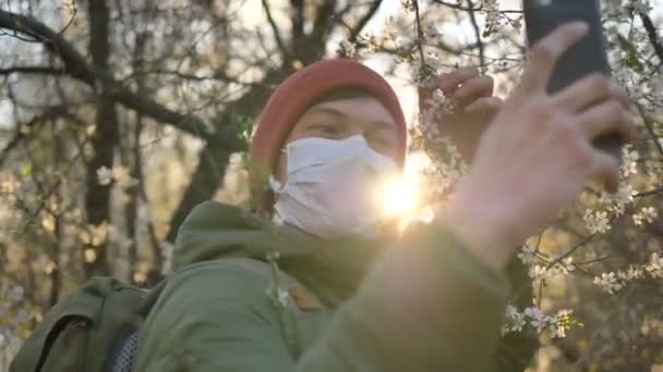 日落时分，戴着医疗面罩的男人拿着一棵盛开的苹果树在电话里自拍 — 图库视频影像