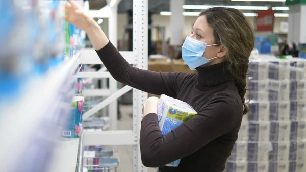 Een jonge vrouw met een medisch masker neemt veel toiletpapier mee in een supermarkt. — Stockfoto