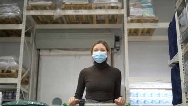 Mujer en máscara médica con carrito de comestibles caminando entre filas en el supermercado — Vídeo de stock