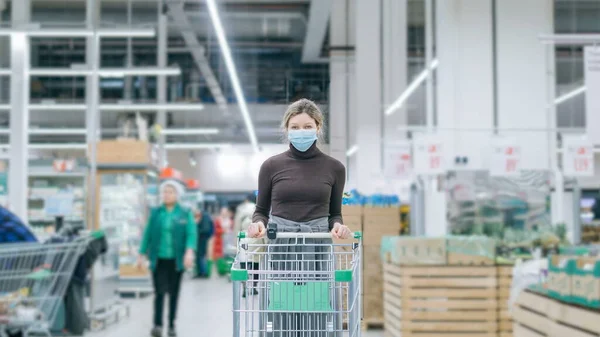 Tıbbi maskeli bir kadın süpermarkette elinde market arabasıyla duruyor. Koronavirüsten koruma, kriz anında yiyecek satın alma..