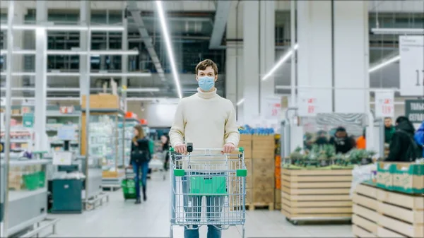 En man i medicinsk mask står med en matvagn i en stormarknad. Skydd mot coronavirus, inköp av viktiga varor. — Stockfoto