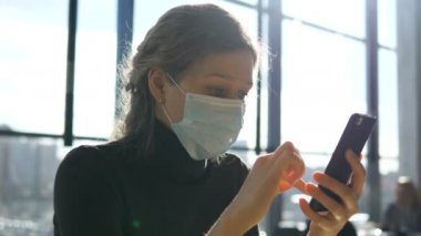 Tıbbi maskeli, ciddi yüzlü bir iş kadını akıllı telefonunun ekranına bakıyor, krizden ve koronavirüsten kaynaklanan mali kayıpları hesaplıyor.