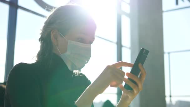 Mladá žena v lékařské masce sedí s telefonem na letišti a čeká na odlet. — Stock video