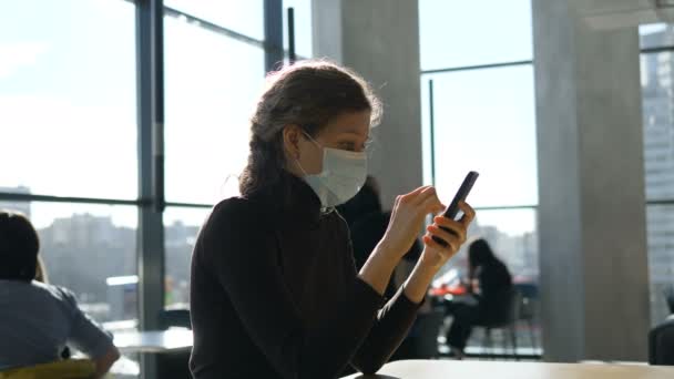 Молодая красивая девушка в медицинской маске сидит с телефоном в торговом центре — стоковое видео