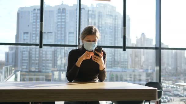Donna d'affari in maschera medica siede con il suo smartphone a un grande tavolo sullo sfondo di grandi finestre panoramiche — Video Stock