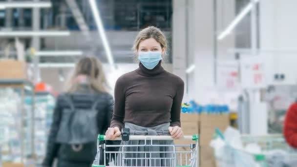 Женщина в медицинской маске стоит в супермаркете с тележкой с продуктами, время истекло. Защита от коронавируса, покупка продуктов питания в условиях кризиса . — стоковое видео